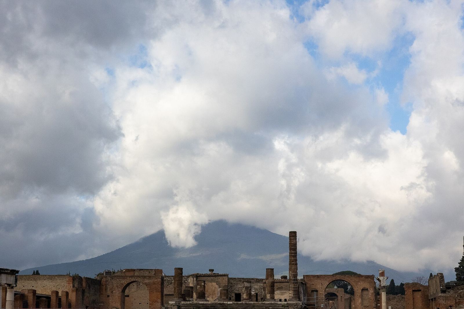 The ancient ruins of Pompeii with the silhouette of Mount Vesuvius rising behind them under a cloudy sky